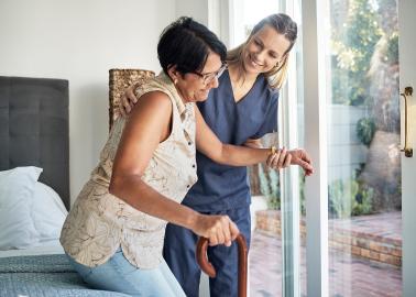A woman in nurses scrubs helps a middle aged woman from a bed, the older woman is holding a cane